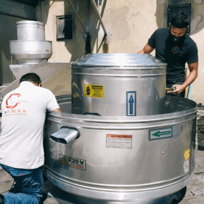 Dos trabajadores inspeccionando un equipo industrial metálico en una planta de procesamiento o fabricación. El equipo tiene etiquetas de advertencia e información.